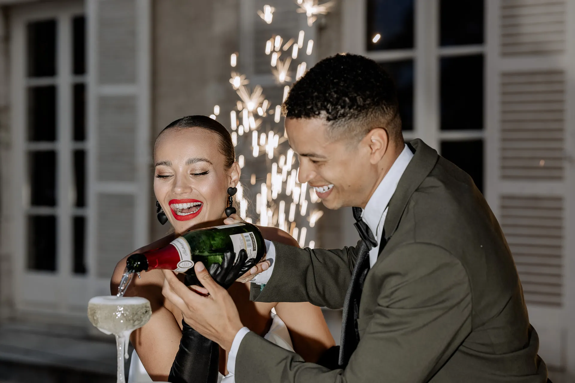 Brautpaar schenkt gemeinsam Champagner ein mit Feuerwerk im Hintergrund am Chateau de Varennes